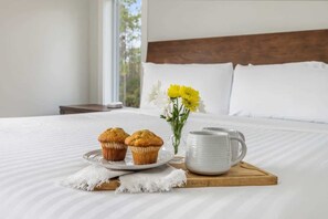 Una cama bien hecha con cabecera de madera, una bandeja con muffins y una taza, y un florero con flores.