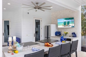 An outdoor patio with a dining table, chairs, a ceiling fan, a TV screen displaying a beach scene, and a kitchen area with a refrigerator and cabinets.