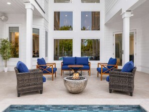 A spacious outdoor seating area with wicker furniture and blue cushions, a central fire pit, and a view of a swimming pool through large windows.