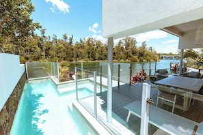 A modern outdoor pool area with glass railings, a wooden deck, and a view of a lake and trees.