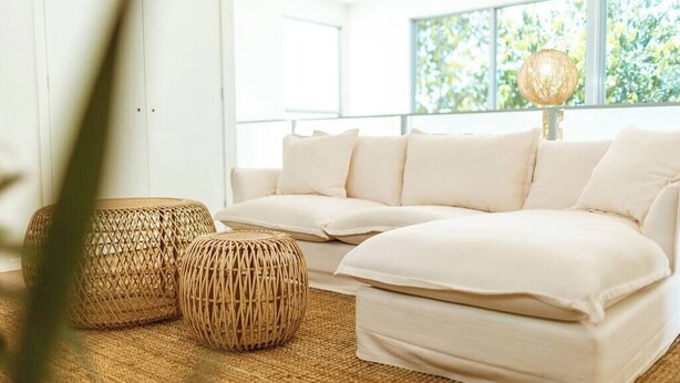 A modern living room with a sectional sofa, two woven ottomans, and a large window with a view of greenery. - Waterfront at Serenity (Maroochydore)