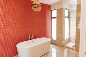 A modern bathroom with a large freestanding bathtub, red mosaic tiles on the walls, and a large window providing natural light.