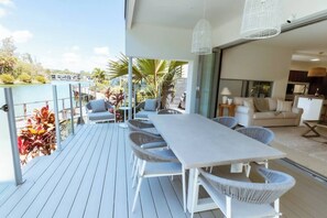 A modern outdoor deck with a dining area, including a white table and wicker chairs, overlooking a body of water with palm trees and a building in the background.