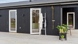 Ein modernes Gebäude mit großen Glasüren, einer weißen Leiter und Topfpflanzen auf einer Holzdielenterrasse.