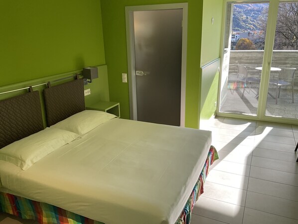 A bed with white linens, a green wall, a door, and a view of a balcony with chairs. - Hotel Liberty Self Check-in (Bellinzona)