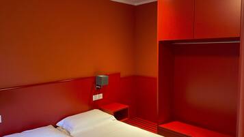 Une chambre d’hôtel avec un lit, une table de chevet, une armoire rouge et une lampe fixée au mur.