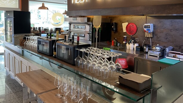 A restaurant with a glass counter displaying wine glasses and bottles, a neon sign reading "PIZZA STEAK GRILL", and a kitchen area with ovens and cooking equipment. - Hotel Liberty Self Check-in (Bellinzona)