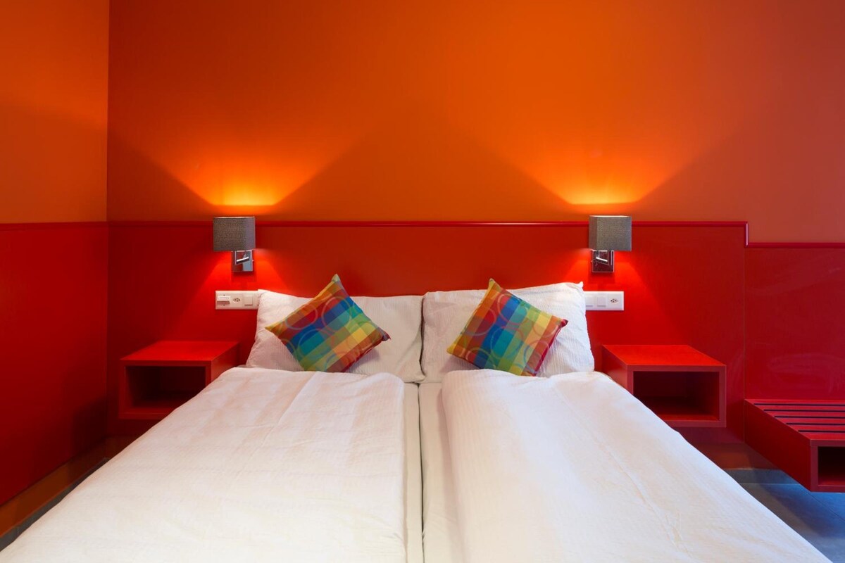 A double bed with white linens and two colorful pillows, flanked by red nightstands with wall-mounted lamps.