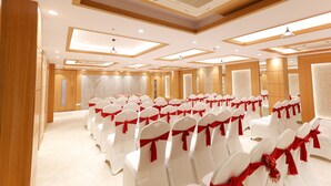A conference room with rows of white chairs, each with a red ribbon. The room has a high ceiling with recessed lighting and wooden paneling. - Hotel Sarc Inn (Varanasi)