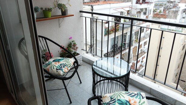 Un balcon avec une table en verre et deux chaises, des plantes en pot et une vue sur le paysage urbain.
