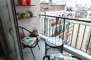 Un balcon avec une table en verre et deux chaises, des plantes en pot et une vue sur le paysage urbain.