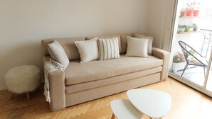 Un canapé beige agrémenté de quatre coussins, un tabouret moelleux blanc et une table blanche dans une pièce dont le sol est en bois, avec une porte-fenêtre coulissante donnant sur un balcon où sont disposées des plantes en pot.