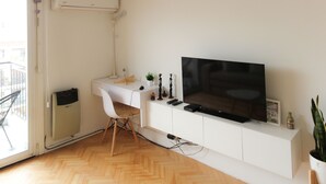 Un salon moderne avec un bureau blanc, un téléviseur à écran plat, une chaise blanche, une petite plante en pot et un radiateur.