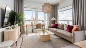 Un salon moderne avec un canapé, une table basse et un téléviseur, de grandes fenêtres offrant une vue sur la ville et un luminaire suspendu.