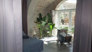 A room with a bed, a lamp, a potted plant, a baby stroller, and a view through a window. - Gites la Remise (PUIMISSON)