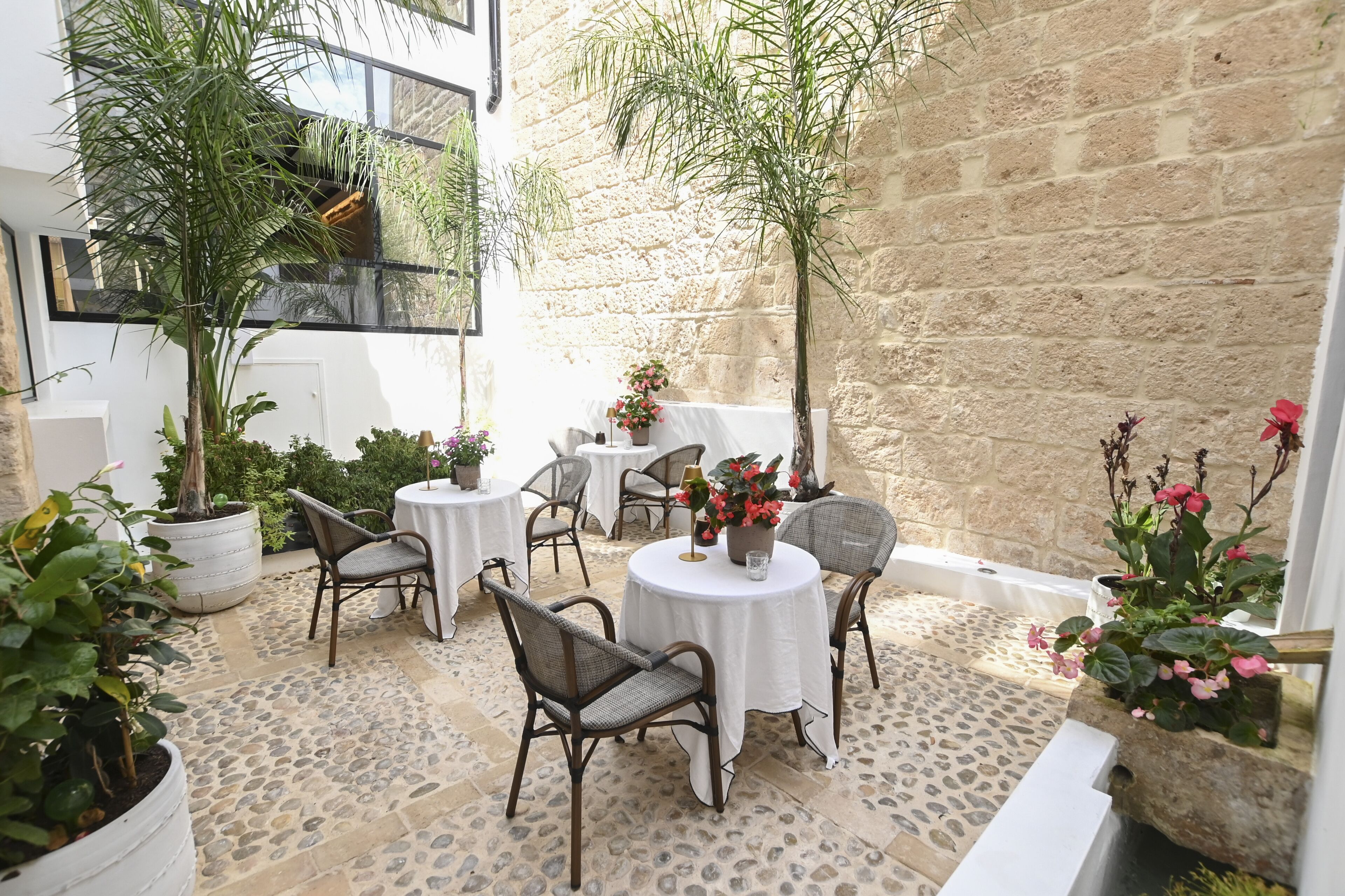 Un patio al aire libre con suelo de piedra, mesas blancas, sillas de mimbre y plantas en macetas.