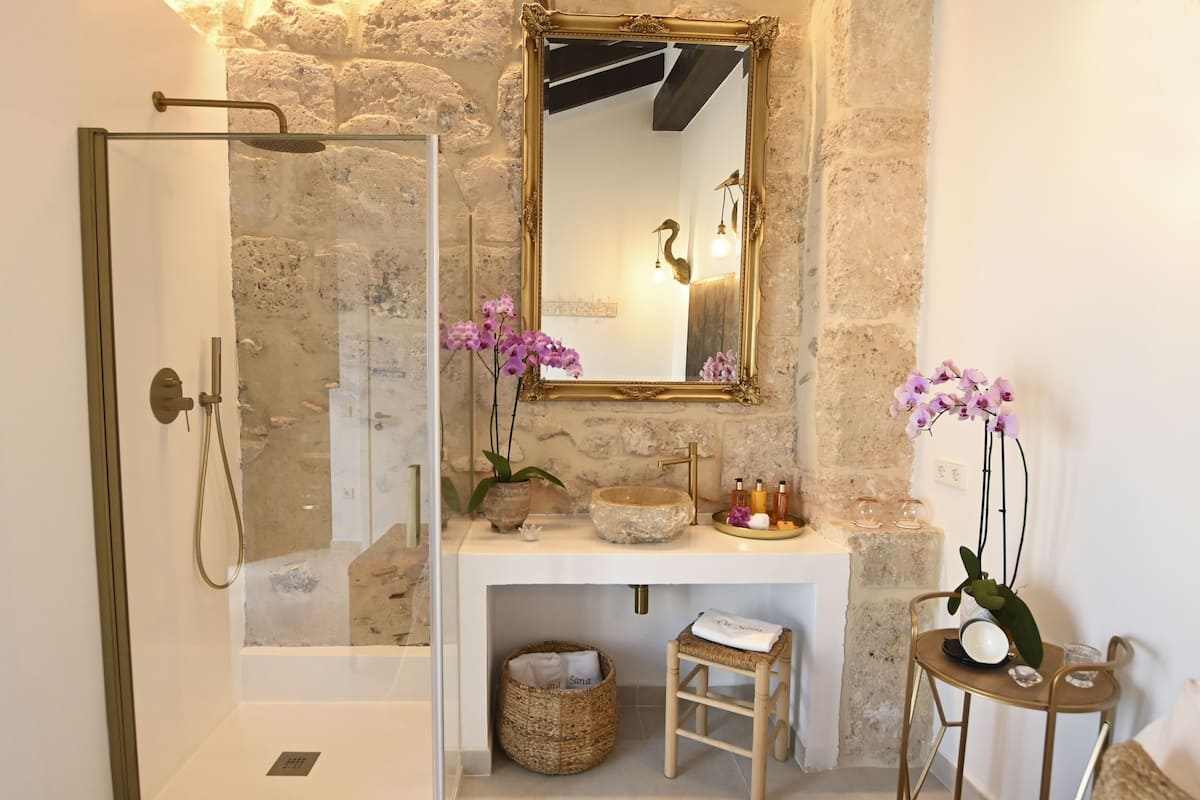 A bathroom with a glass shower enclosure, a stone sink, and a mirror with a gold frame.