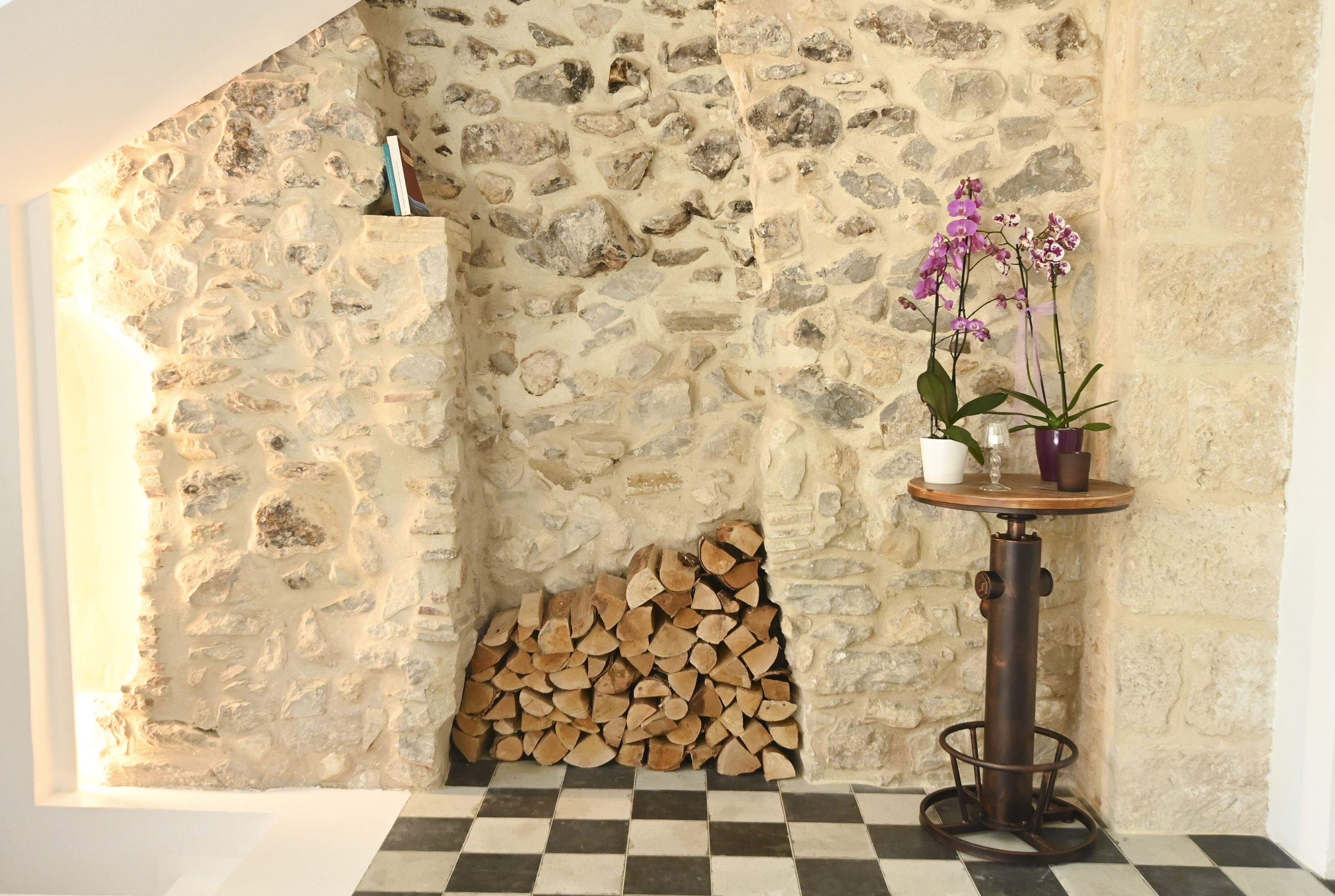 Un mur de pierres avec une cheminée, une pile de bois de chauffage et une table ornée de pots d’orchidées.