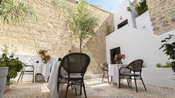 Une terrasse extérieure avec un sol en pierre, des tables blanches, des chaises en osier et des plantes en pot.