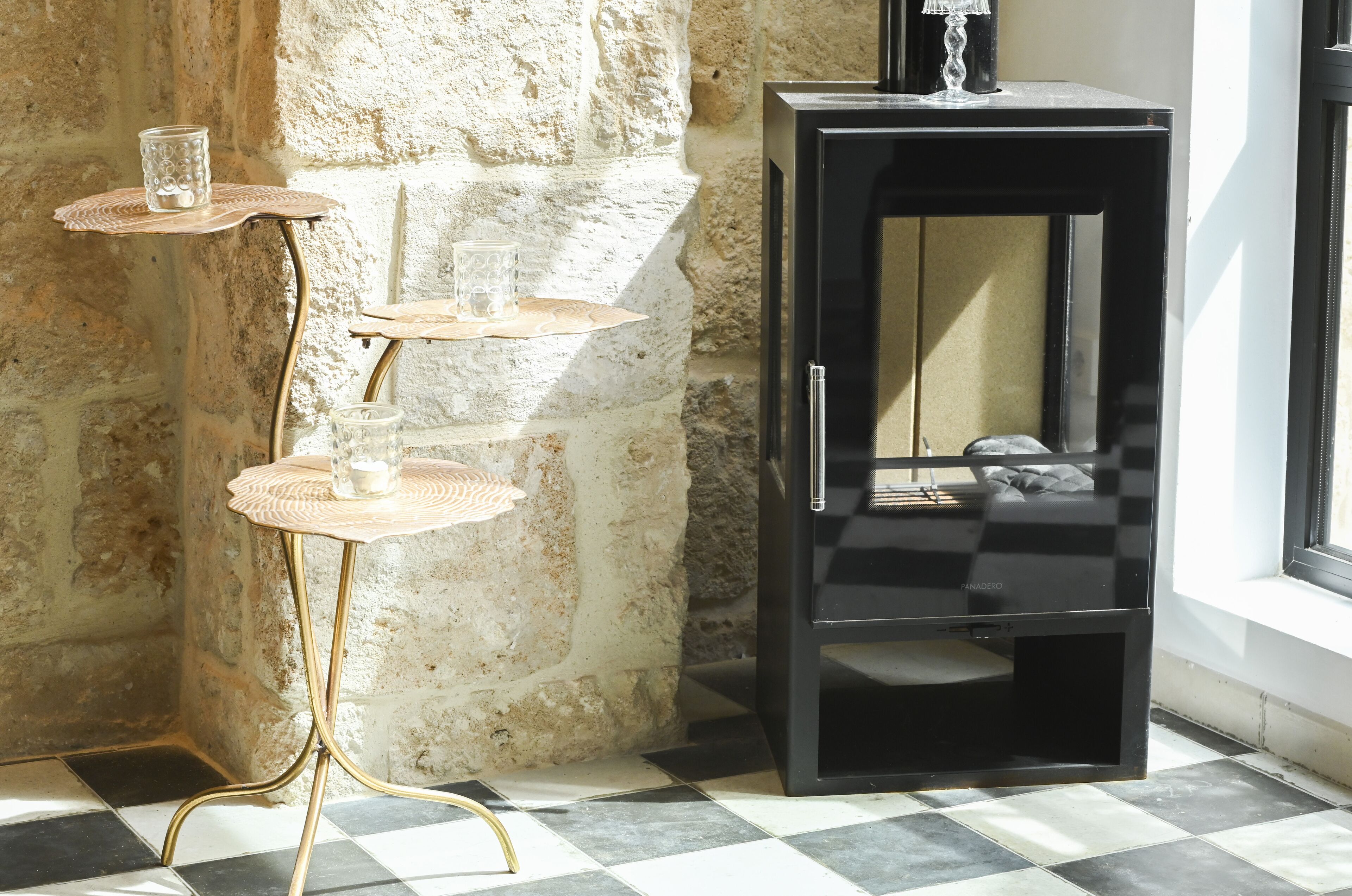 Un coin aménagé avec une table à trois niveaux où sont disposés des verres, un meuble noir et un mur en pierre.