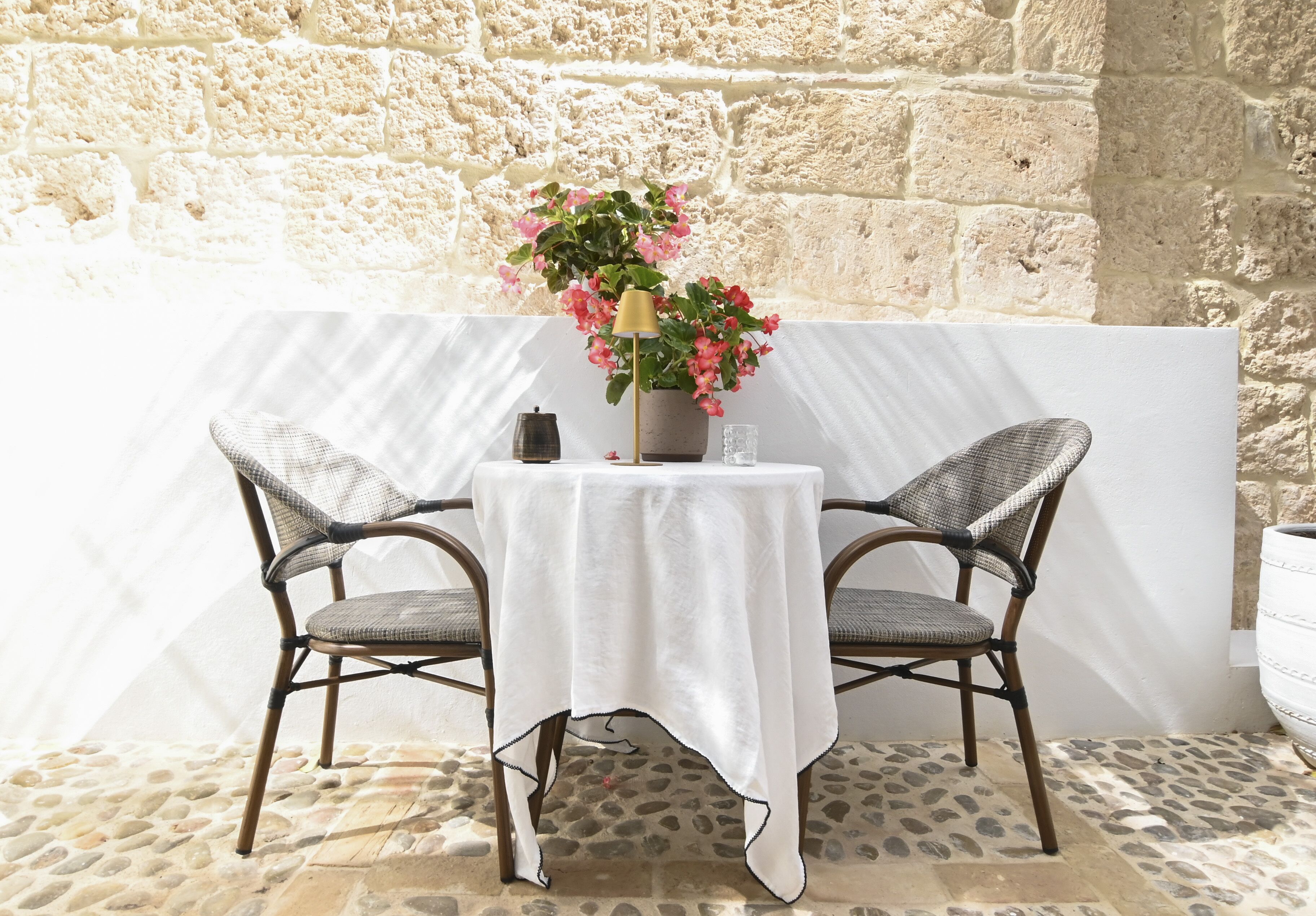 A table with a white tablecloth, two wicker chairs, a potted plant with pink flowers, and a bell on the table.