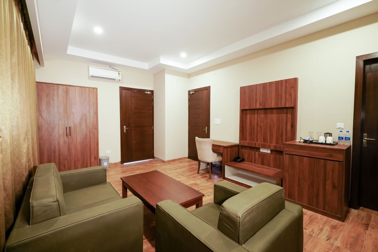 A modern living room with a sofa, coffee table, and a desk area. There are wooden wardrobes and a small kitchenette with a sink and countertop.