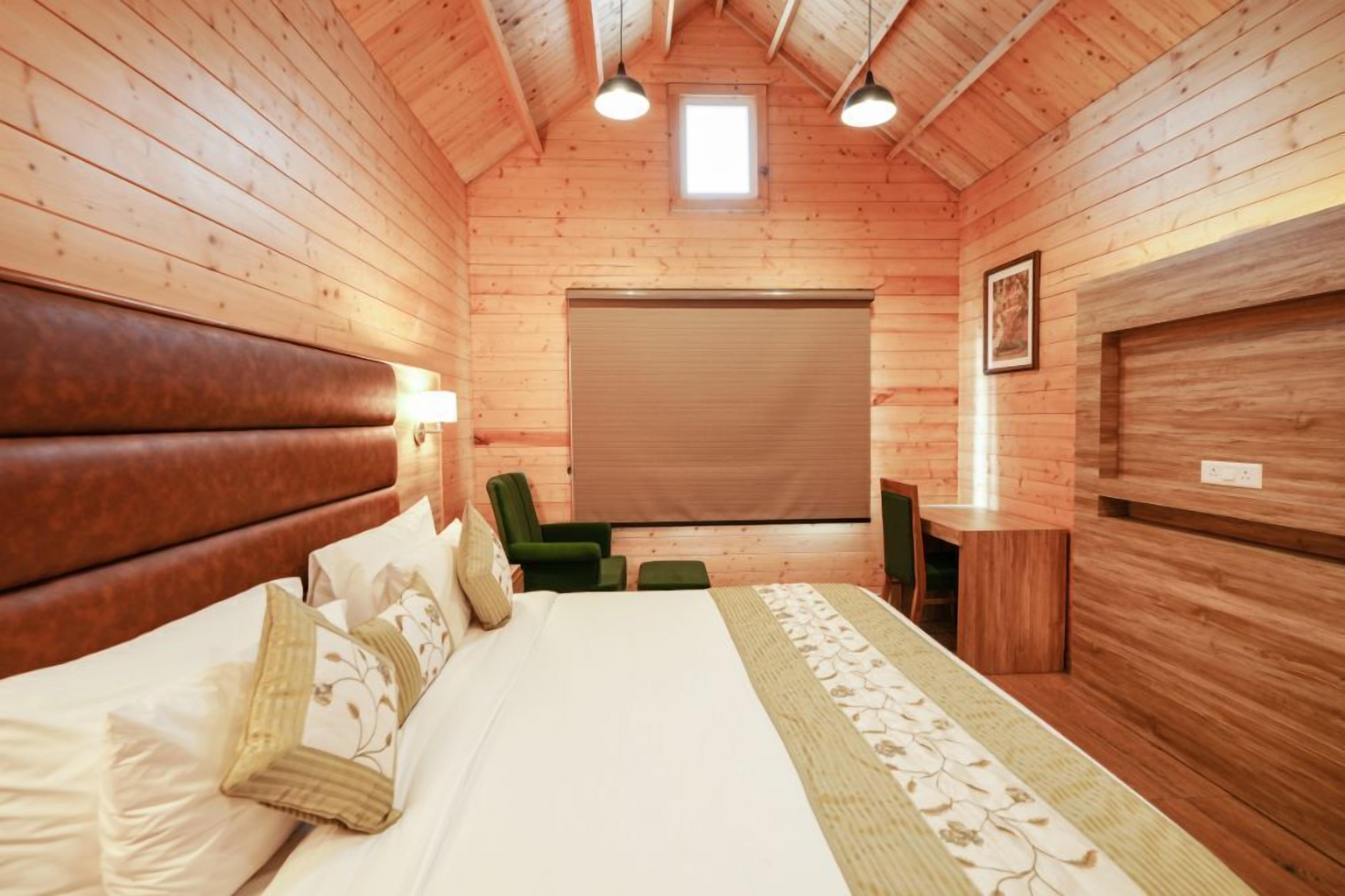 A wooden cabin room with a bed, a chair, a desk, and a window.