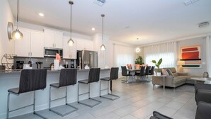 Eine moderne Küche mit weißen Schränken, Edelstahlgeräten und Barhocker. Es gibt einen Essbereich mit Tisch und Stühlen sowie ein Wohnzimmer mit Sofa und Couchtisch.