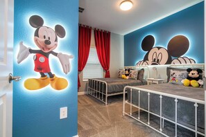 Una habitación infantil con decoración de Mickey Mouse, una cama con estructura de metal y una ventana con cortinas rojas.
