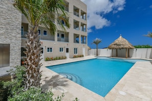 A swimming pool with a stone wall, palm trees, and a thatched-roof structure. - Aruba Coco het Tropische Luxe Verblijf met een Fantastische Centrale Ligging (Oranjestad)