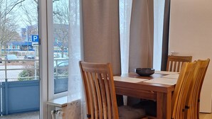 Ruokailutilassa on puinen pöytä ja tuolit, valkotaulu sekä suuret ikkunat, joista on näkymä ulos, ja ikkunoissa on verhot.