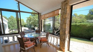 Une terrasse ensoleillée avec une table ronde et des chaises, de grandes fenêtres et un pilier en pierre.