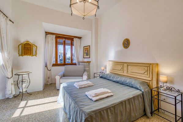 A bedroom with a bed, a chair, a table, and a window with curtains. - Chianti Classico Home Panoramic View (Castellina in Chianti)