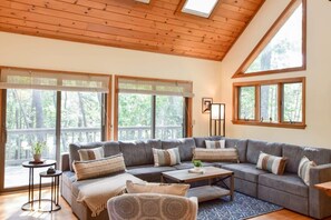A cozy living room with a sectional sofa, a coffee table, and a small side table. There are large windows with blinds, and a skylight above the ceiling. - 4 Mins to Wellfleet Village, Screened sunroom! (Wellfleet)