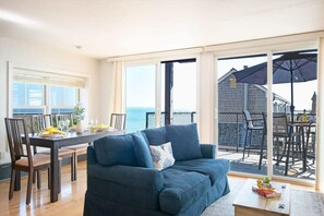 A modern living room with a sofa, dining table, and chairs. There is a large sliding glass door leading to an outdoor patio with a table and chairs. - Gorgeous water views in the center of town! (Provincetown)