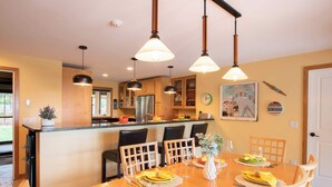 A modern kitchen with wooden cabinets, a stainless steel refrigerator, and a dining area set for a meal with a table, chairs, and a decorative wall clock. - Beautiful backyard, pool, deeded beach rights! (Truro)