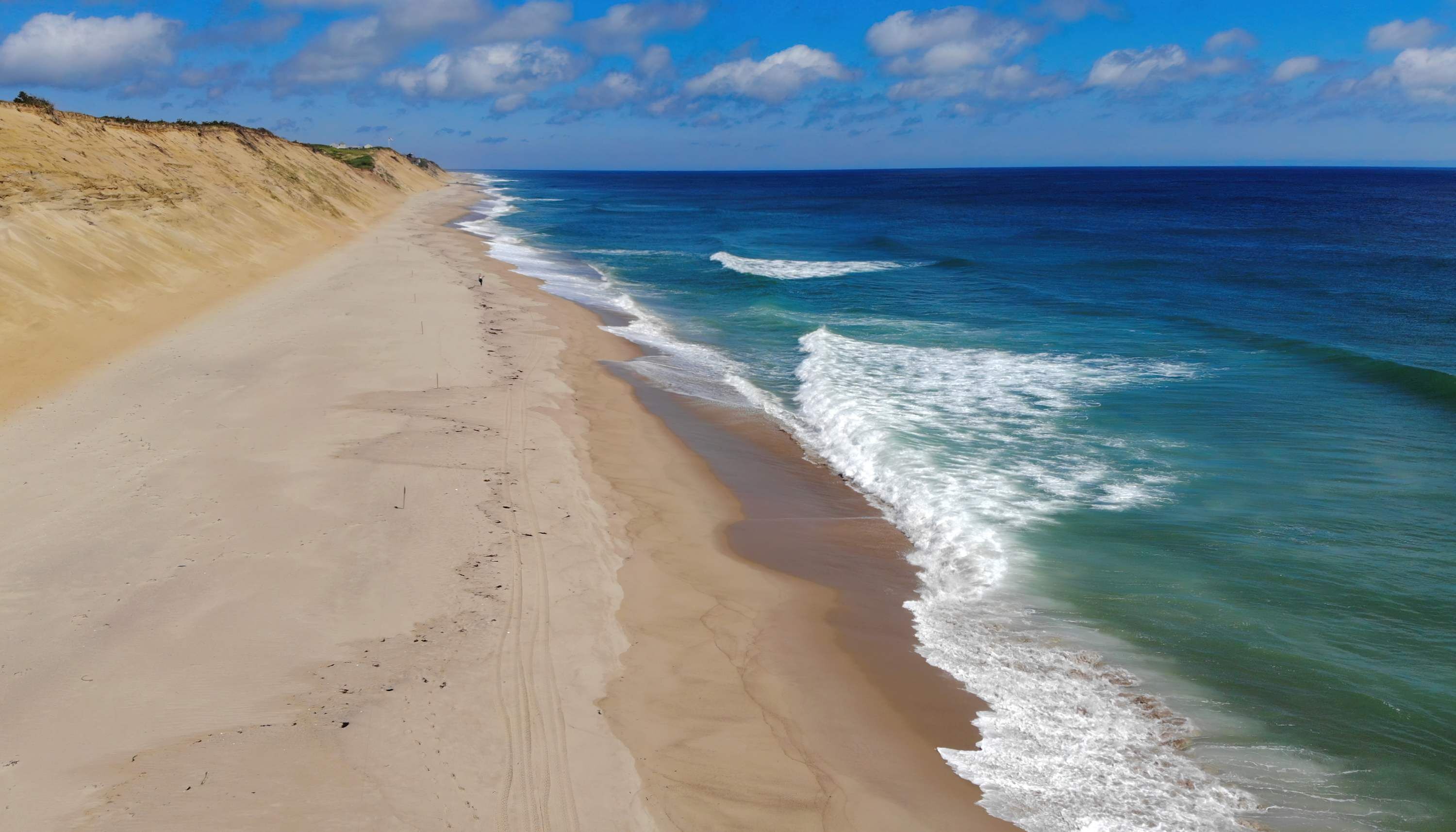 A wide sandy beach with gentle waves, a clear sky, and a distant cliff.