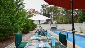 An outdoor dining area with a glass table, chairs, and a pool in the background. - Beautiful backyard, pool, deeded beach rights! (Truro)