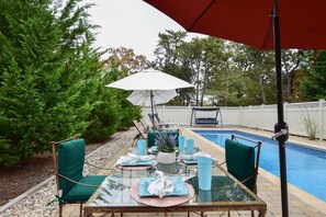 Ein Esstisch mit Glasplatte, Stühle, ein weißer Sonnenschirm und ein Pool im Hintergrund.