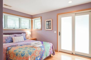 Un dormitorio con cama, mesita de noche, lámpara, ventana con persianas, puerta y un cuadro colgado en la pared.