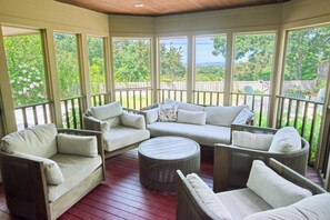 Un espacio exterior techado con zona de estar de madera, asientos acolchados y ventanas amplias con vistas al verdor.