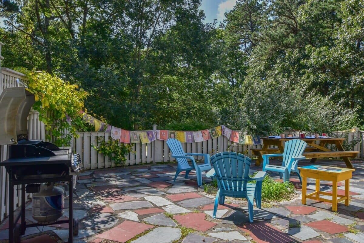 An outdoor patio with stone flooring, blue Adirondack chairs, and a barbecue grill.