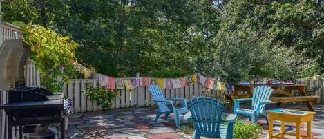 An outdoor patio with stone flooring, blue Adirondack chairs, and a barbecue grill.