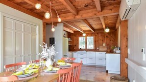 Un comedor con una mesa de madera dispuesta para cuatro personas, rodeada de sillas a juego. Se ve una cocina al fondo con armarios blancos y una encimera de madera.