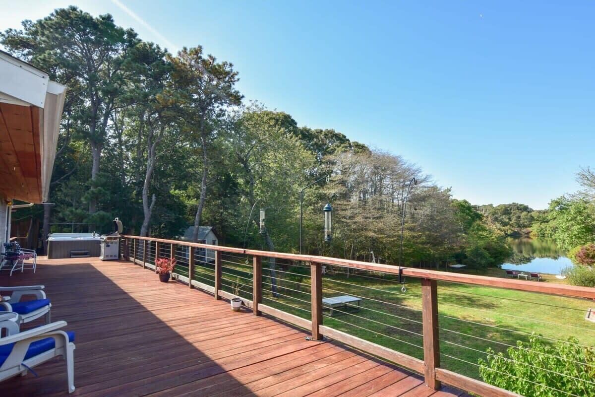 A wooden deck with a railing, outdoor seating, and a view of a lake surrounded by trees.