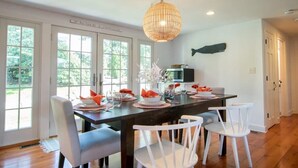 A dining area with a dark wooden table, white chairs, and a large woven pendant light. - Roof Deck, Walk to Forest Beach, Fire Pit! (Chatham)