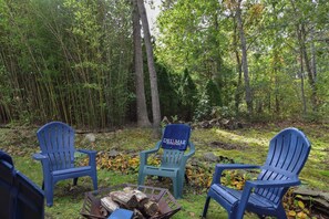 Three blue Adirondack chairs arranged around a small fire pit in a forested area with a sign reading 'DEL MAR Vacation Rentals.'