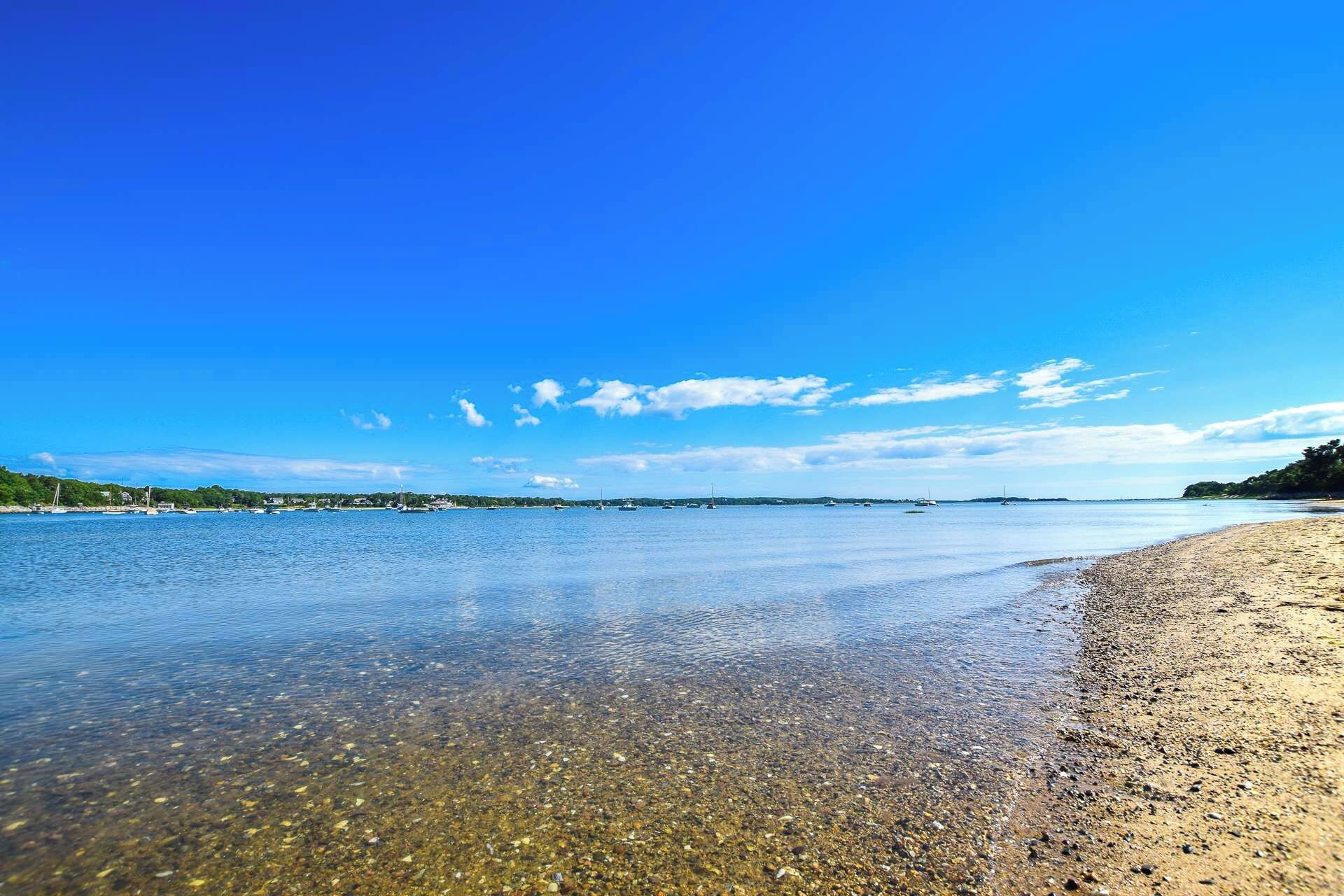 Una playa con agua cristalina, piedras y barcos a lo lejos.