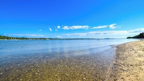 Una playa con agua cristalina, piedras y barcos a lo lejos.