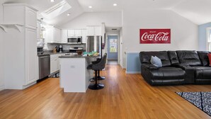 A modern kitchen with white cabinets, stainless steel appliances, and a central island with bar stools. A black leather couch and a Coca-Cola sign are in the living area. - Hot Tub, 200 ft to Beach! (Dennis)